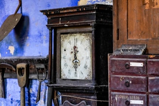 A vintage, ornate clock with roman numerals is set against a rustic backdrop. On one side, wooden tools hang against a worn blue wall, while on the other, a wooden cabinet with labeled drawers sits. The overall setting suggests an old workshop or a quaint antique space.