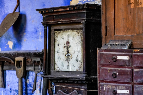 A vintage, ornate clock with roman numerals is set against a rustic backdrop. On one side, wooden tools hang against a worn blue wall, while on the other, a wooden cabinet with labeled drawers sits. The overall setting suggests an old workshop or a quaint antique space.