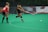 Two athletes are playing field hockey on a green artificial turf. One player, in a black uniform, is actively striking a white ball with a pink stick, while the other player, wearing a red uniform, watches attentively.