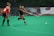 Two athletes are playing field hockey on a green artificial turf. One player, in a black uniform, is actively striking a white ball with a pink stick, while the other player, wearing a red uniform, watches attentively.