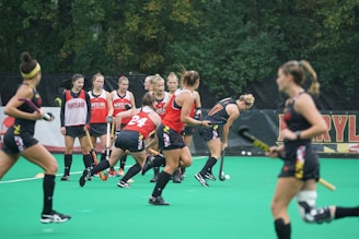 A group of athletes engaged in a sports practice session on a green field. They are wearing red and black athletic uniforms, some with the word 'Maryland' visible. Field hockey sticks are held by the players, and there is a sense of movement and activity as they appear to be in play. Trees in the background suggest an outdoor setting.