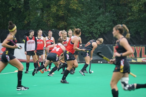 A group of athletes engaged in a sports practice session on a green field. They are wearing red and black athletic uniforms, some with the word 'Maryland' visible. Field hockey sticks are held by the players, and there is a sense of movement and activity as they appear to be in play. Trees in the background suggest an outdoor setting.