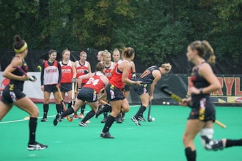 A group of athletes engaged in a sports practice session on a green field. They are wearing red and black athletic uniforms, some with the word 'Maryland' visible. Field hockey sticks are held by the players, and there is a sense of movement and activity as they appear to be in play. Trees in the background suggest an outdoor setting.