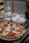 A delicious-looking pizza topped with pepperoni, green peppers, and melted mozzarella cheese sits on a wooden table. Next to it is a clear glass bottle of water, a stack of plates, and some cutlery wrapped in a napkin.