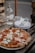 A delicious-looking pizza topped with pepperoni, green peppers, and melted mozzarella cheese sits on a wooden table. Next to it is a clear glass bottle of water, a stack of plates, and some cutlery wrapped in a napkin.