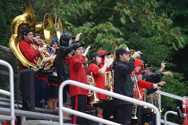A group of people wearing raincoats and jackets, holding brass instruments, stand in a row on bleachers. They appear to be part of a marching band, preparing or performing in an outdoor setting. The weather looks rainy, as suggested by their attire. Lush green trees are visible in the background.