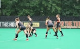 Several athletes in matching sports uniforms are on a bright green field, engaged in a field hockey game. They appear focused and are holding hockey sticks, with some bending slightly as they prepare to play. Trees and a billboard are visible in the background.