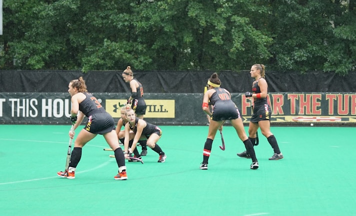 Several athletes in matching sports uniforms are on a bright green field, engaged in a field hockey game. They appear focused and are holding hockey sticks, with some bending slightly as they prepare to play. Trees and a billboard are visible in the background.
