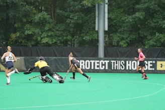 A field hockey match in progress with players actively engaging in the game. One player in yellow appears to be a goalkeeper attempting to block or stop the play, while two outfield players are chasing after the ball. A referee in a pink top runs nearby. The background includes lush green trees and a banner with a slogan.