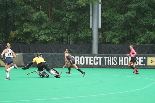 A field hockey match in progress with players actively engaging in the game. One player in yellow appears to be a goalkeeper attempting to block or stop the play, while two outfield players are chasing after the ball. A referee in a pink top runs nearby. The background includes lush green trees and a banner with a slogan.
