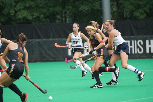 A group of female athletes are playing field hockey on a green turf field. They are wearing uniforms, with two distinct sets of colors representing different teams. The players are actively engaged, with focus on the ball that is in play on the ground.