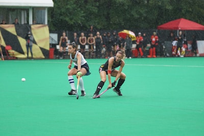 Two field hockey players are engaged in a match on a wet, green field. One player in a white and navy uniform is closely following another player in a black uniform who is controlling the ball with a hockey stick. Spectators and team members are visible in the background, some under a red tent.