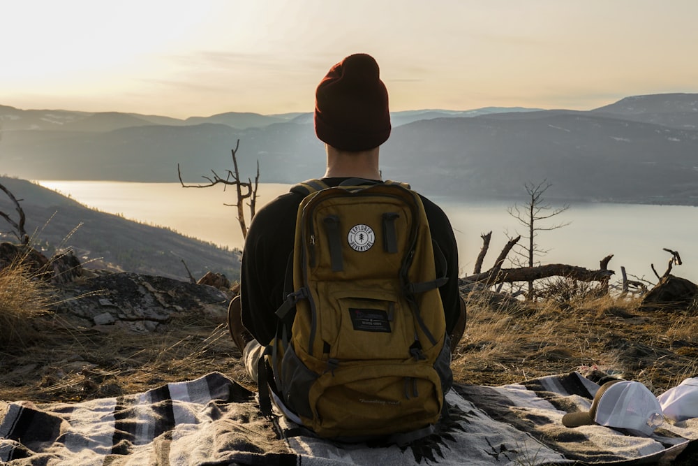 person sitting on mat carrying backpack while looking on the lake