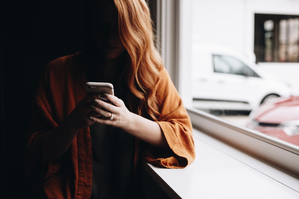 woman leaning beside window while using iPhone