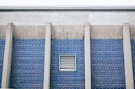 A section of a building wall with blue brick tiles and vertical concrete columns. A rectangular ventilation grille is set in the middle of the brick wall.
