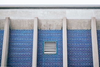 A section of a building wall with blue brick tiles and vertical concrete columns. A rectangular ventilation grille is set in the middle of the brick wall.