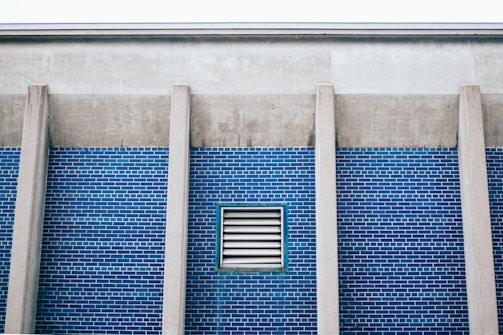 A section of a building wall with blue brick tiles and vertical concrete columns. A rectangular ventilation grille is set in the middle of the brick wall.