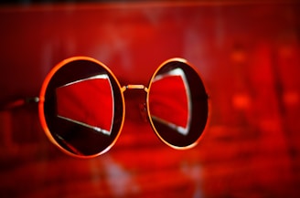 A pair of round sunglasses with gold frames against an intense red background, with reflections creating geometric patterns on the lenses.
