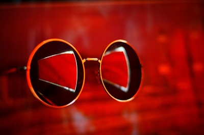 A pair of round sunglasses with gold frames against an intense red background, with reflections creating geometric patterns on the lenses.