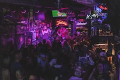 A lively bar scene with a crowd gathered around a stage where a band is performing. Neon signs and dim lighting create a vibrant atmosphere. The room is bustling with people enjoying drinks and socializing.
