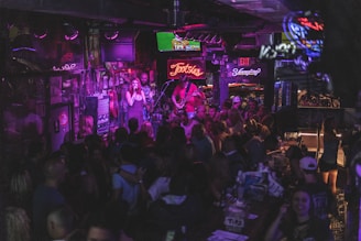 A lively bar scene with a crowd gathered around a stage where a band is performing. Neon signs and dim lighting create a vibrant atmosphere. The room is bustling with people enjoying drinks and socializing.