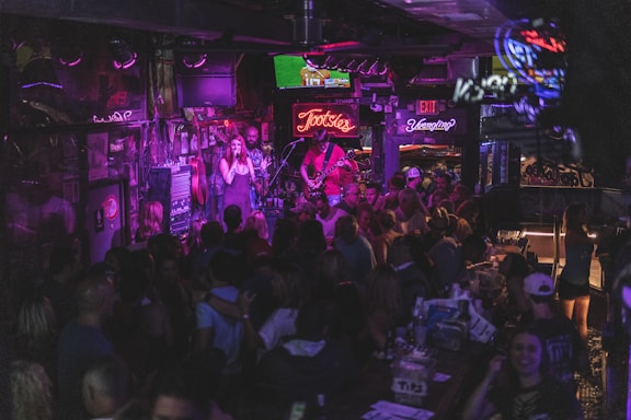 A lively bar scene with a crowd gathered around a stage where a band is performing. Neon signs and dim lighting create a vibrant atmosphere. The room is bustling with people enjoying drinks and socializing.