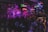 A lively bar scene with a crowd gathered around a stage where a band is performing. Neon signs and dim lighting create a vibrant atmosphere. The room is bustling with people enjoying drinks and socializing.
