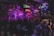 A lively bar scene with a crowd gathered around a stage where a band is performing. Neon signs and dim lighting create a vibrant atmosphere. The room is bustling with people enjoying drinks and socializing.