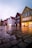 A street scene features a row of colorful wooden buildings with steep roofs, typical of Scandinavian architecture. The cobblestone pavement is wet, reflecting the buildings in large puddles. Warm lights in the windows contrast with the cool, overcast sky.