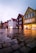 A street scene features a row of colorful wooden buildings with steep roofs, typical of Scandinavian architecture. The cobblestone pavement is wet, reflecting the buildings in large puddles. Warm lights in the windows contrast with the cool, overcast sky.
