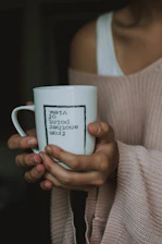 A person holds a white mug with both hands. The mug features the text 'From another point of view' printed upside down. The individual is wearing a soft pink knitted sweater, and the background is a dark, blurred interior.