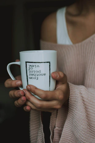 A person holds a white mug with both hands. The mug features the text 'From another point of view' printed upside down. The individual is wearing a soft pink knitted sweater, and the background is a dark, blurred interior.