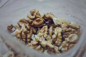 A close-up view of several walnuts inside a transparent glass container. The walnuts are shelled, revealing their intricate patterns and textures. The focus is on the nuts, with a blurred background giving a soft appearance.