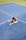 A person is sitting on the ground of a tennis court with legs stretched and head bowed down. The court surface is blue with white boundary lines, and a patch of green can be seen in the background.