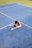 A person is sitting on the ground of a tennis court with legs stretched and head bowed down. The court surface is blue with white boundary lines, and a patch of green can be seen in the background.