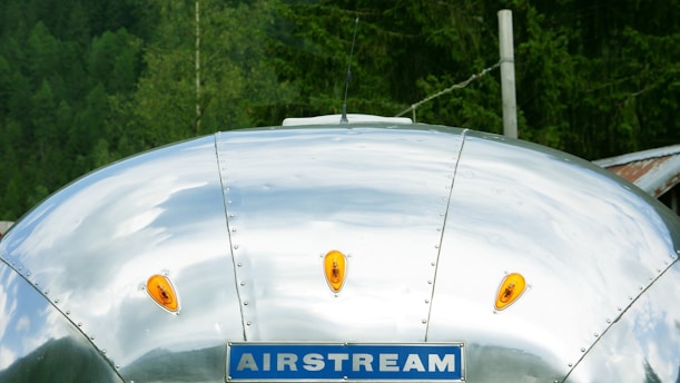 A silver metallic surface with rivets visible, featuring orange reflector lights and a blue sign with the word 'AIRSTREAM.' The background includes greenery, suggesting a forest setting.