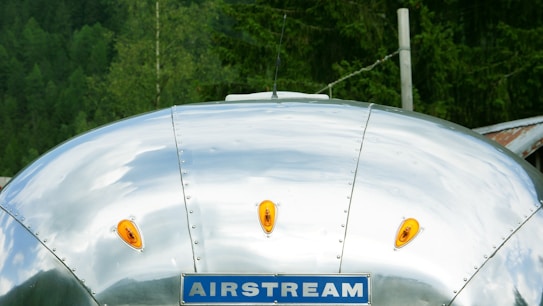 A silver metallic surface with rivets visible, featuring orange reflector lights and a blue sign with the word 'AIRSTREAM.' The background includes greenery, suggesting a forest setting.
