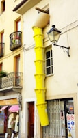 A tall stack of yellow construction waste chutes is attached to the exterior of a building, descending from a higher floor window. The building has wrought iron balconies with flower pots and a street lamp mounted on the wall. There is a clothing store at the street level.