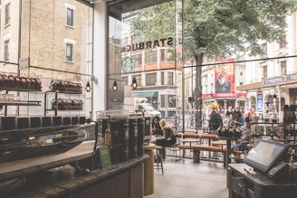 The interior of a busy cafe with large glass windows overlooking a street. Inside, a counter displays various coffee products and machines, while customers sit at tables, engaged in conversation or using their devices. Outside, pedestrians walk past, and bicycles are parked nearby. The scene includes urban structures and signage visible through the window.