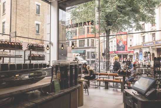 The interior of a busy cafe with large glass windows overlooking a street. Inside, a counter displays various coffee products and machines, while customers sit at tables, engaged in conversation or using their devices. Outside, pedestrians walk past, and bicycles are parked nearby. The scene includes urban structures and signage visible through the window.