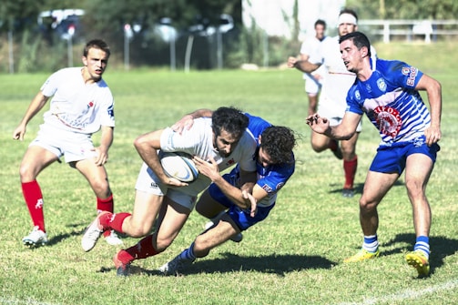 Several rugby players in action on a grassy field, with one player in white attempting to carry the ball while being tackled by a player in blue. Other players in white and blue are visible in the background, actively participating in the match.