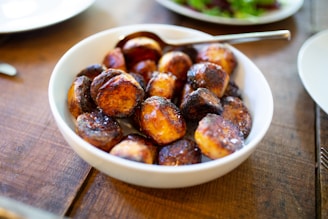 A bowl of roasted potatoes with a crispy, golden-brown exterior served in a white dish. The potatoes appear freshly cooked and are accompanied by a spoon in the dish. The image is taken on a wooden table with part of another plate visible in the background.