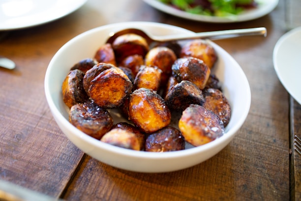 A bowl of roasted potatoes with a crispy, golden-brown exterior served in a white dish. The potatoes appear freshly cooked and are accompanied by a spoon in the dish. The image is taken on a wooden table with part of another plate visible in the background.