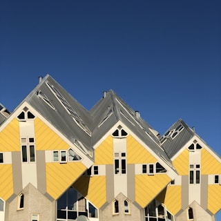 A series of uniquely designed cube-shaped houses with slanted walls and roofs in shades of yellow and grey. The architectural style is modern and abstract, set against a clear blue sky.