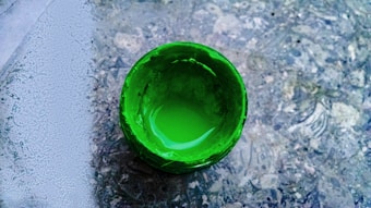 A top-down view of a vibrant green paint jar on a textured surface. The paint appears liquid and glossy, with light reflections on the smooth surface.