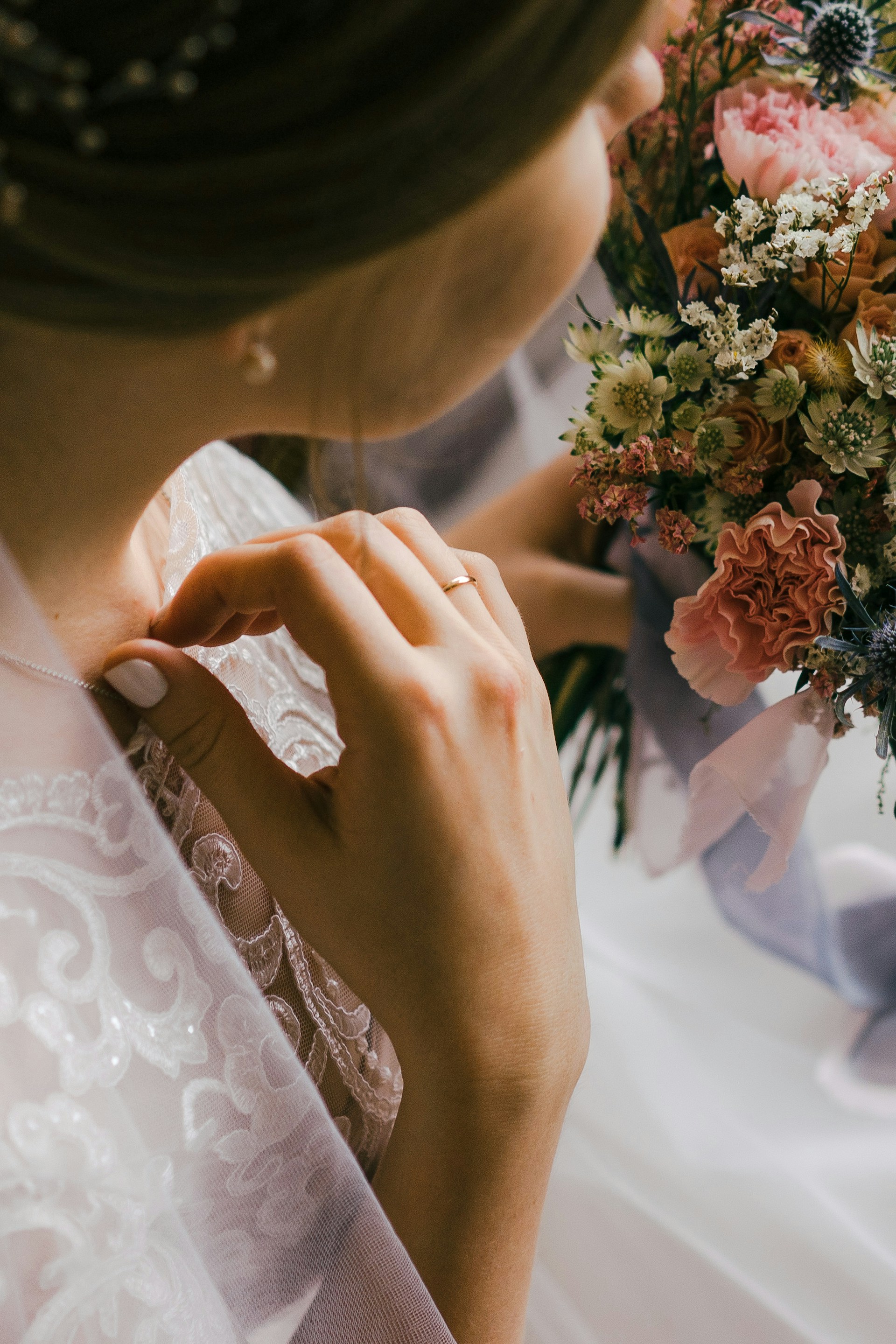 A close-up of a bride's hand touching her lace wedding dress, holding a bouquet filled with pink and white flowers. The focus is on the intricate lace details of the dress and the delicate arrangement of the flowers.