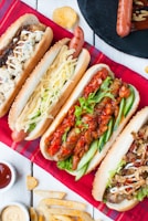 An arrangement of four different types of hot dogs placed on a red plaid cloth, each featuring various toppings like cheese, lettuce, grilled onions, and tomato sauce. Next to the hot dogs, there are small bowls of sauces and some scattered potato chips and fries.