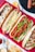 An arrangement of four different types of hot dogs placed on a red plaid cloth, each featuring various toppings like cheese, lettuce, grilled onions, and tomato sauce. Next to the hot dogs, there are small bowls of sauces and some scattered potato chips and fries.
