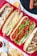 An arrangement of four different types of hot dogs placed on a red plaid cloth, each featuring various toppings like cheese, lettuce, grilled onions, and tomato sauce. Next to the hot dogs, there are small bowls of sauces and some scattered potato chips and fries.