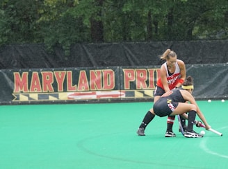 Two female field hockey players, one wearing a red jersey and the other wearing a black jersey with yellow and red accents, are engaged in a play on a green field. They are both focused on controlling the ball, with one player bending down with her stick while the other stands close behind. A banner in the background displays the words 'Maryland Pride'. Trees form the backdrop beyond the banner.